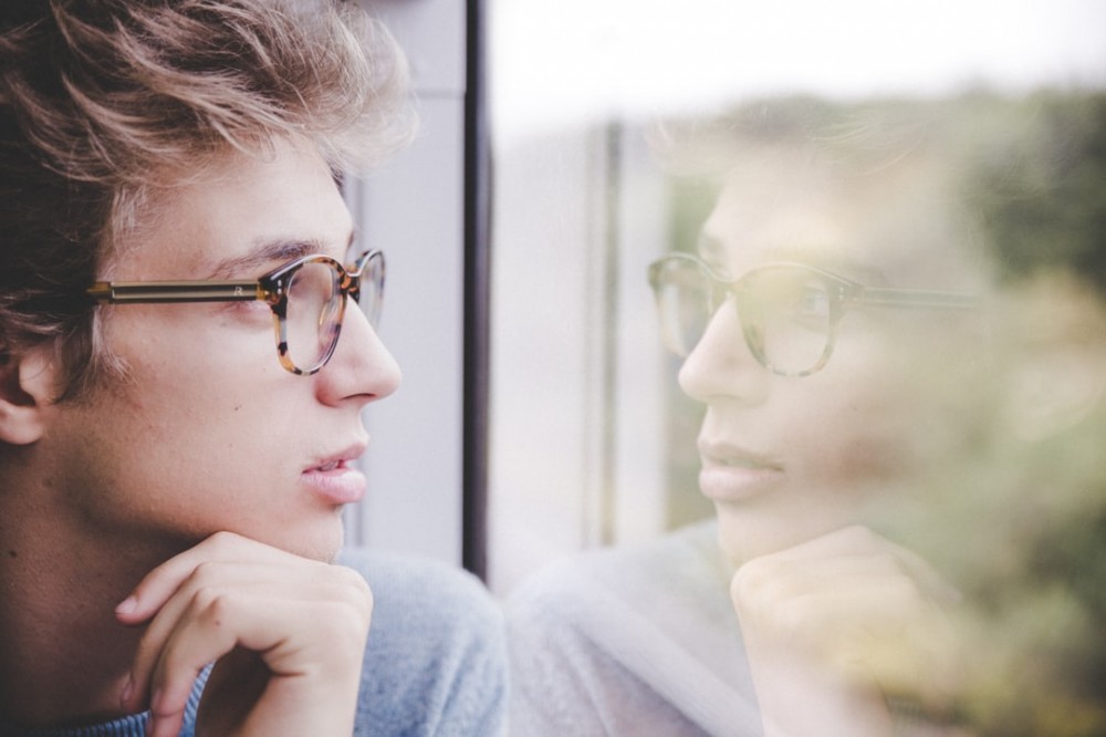 A man with glasses seeing his own reflection in a window